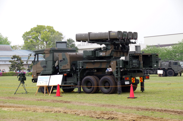 Ein Militärlastwagen mit einer Rakete auf dem Dach ist auf einem Feld geparkt, umgeben von Verkehrskegeln, einem Schild, einer Kamera auf einem Stativ, Menschen, Blumenpflanzen, Bäumen, Gebäuden und einem klaren blauen Himmel.