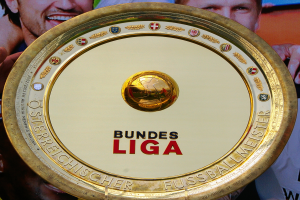 Mann hält goldenen Schild mit 'Bundesliga' hoch, im Hintergrund eine Fahne mit drei Personen
