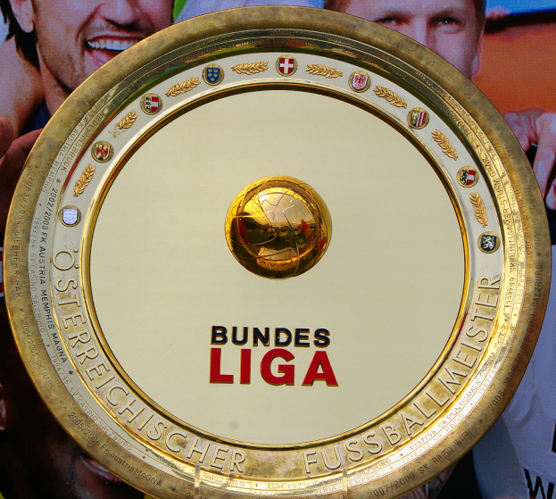 Mann hält goldenen Schild mit 'Bundesliga' hoch, im Hintergrund eine Fahne mit drei Personen