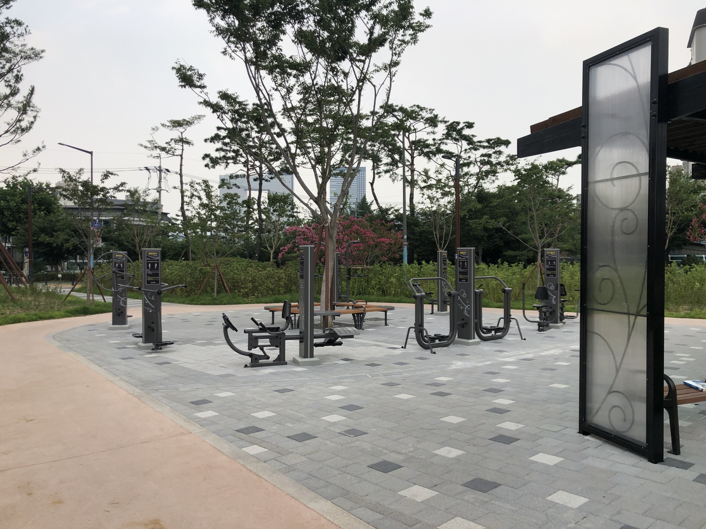 Outdoor-Park mit Fitnessgeräten, Bänken, Bäumen, Pflanzen, Gras, Pfählen, Lampen, Drähten und Gebäuden vor einem Himmel-Hintergrund.