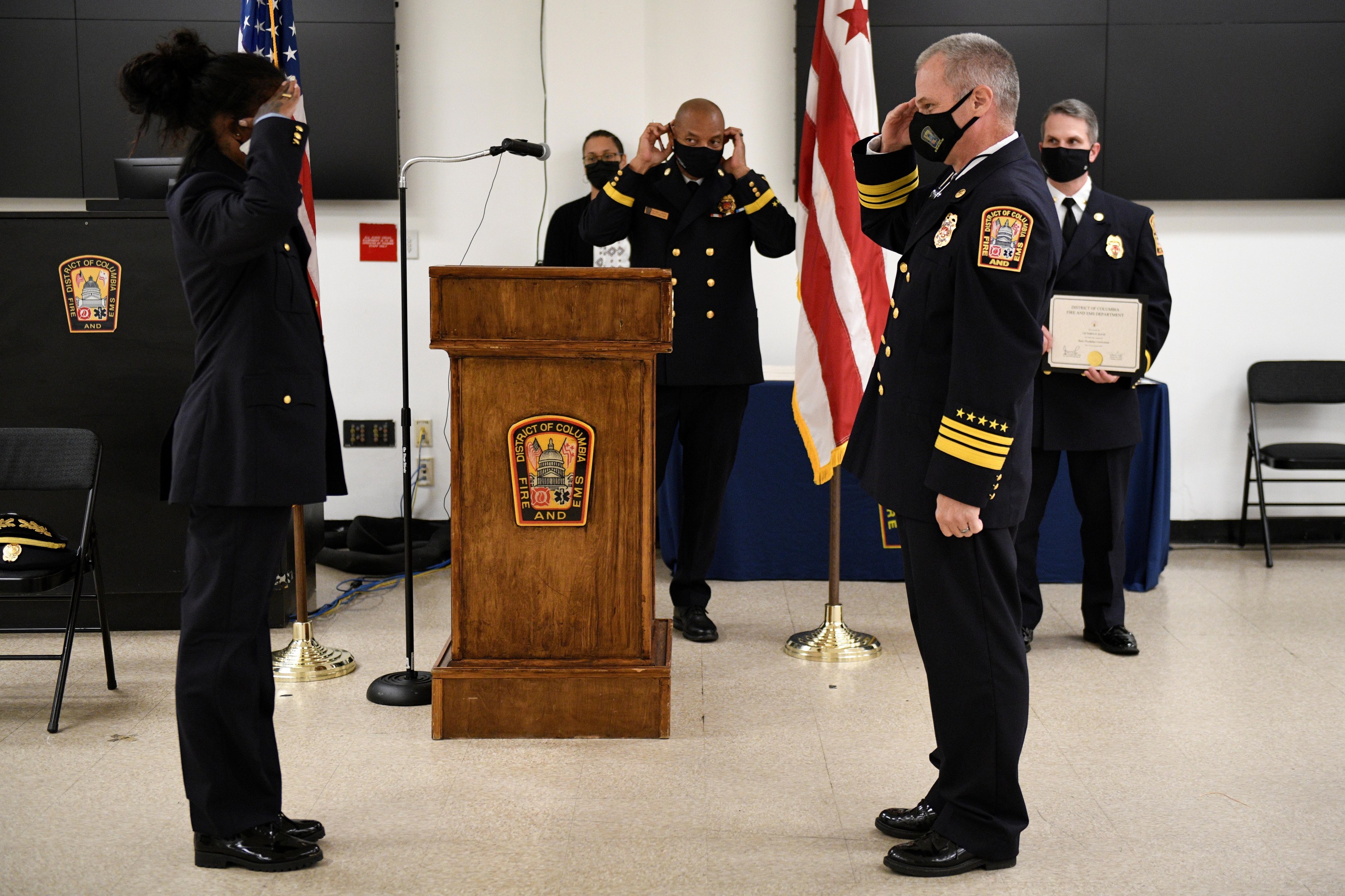 Gruppe uniformierter Personen auf einem Podium während einer Vereidigung eines neuen Feuerchefs, der eine Urkunde hält, mit Flaggen, Stühlen, Tischen und einer Wand im Hintergrund.