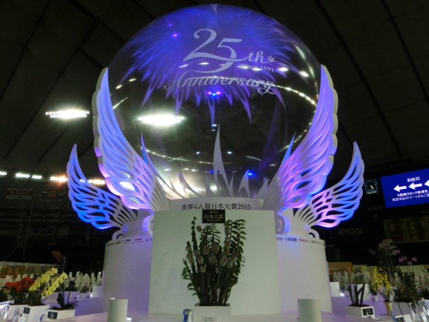 Ein großer Kristallball mit Flügeln oben drauf, umgeben von Blumenbouquets, Namensschildern und anderen Gegenständen, vor einem festlichen Hintergrund mit Schildern, Lichtern und einem Bildschirm für ein 25-jähriges Jubiläum.