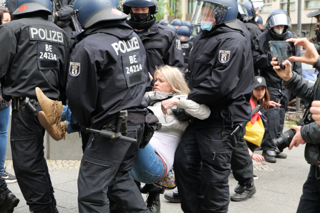 Eine Gruppe von Polizisten in Einsatzausrüstung eskortiert eine Frau in einem weißen und blauen Kleid, wobei ein Polizist ihren Arm hält, während eine andere Person die Szene fotografiert; Gebäude, Bäume und der Himmel sind im Hintergrund sichtbar.