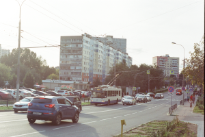 Eine belebte Stadtstraße mit zahlreichen Autos, Bussen, Straßenlaternen, Schildern, Laternenmasten, Strommasten mit Drähten, Bäumen und Gebäuden unter einem klaren blauen Himmel.