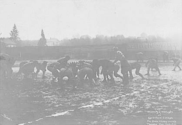 Ein Schwarz-Weiß-Foto einer Gruppe von Menschen, die Fußball auf einem Feld spielen, mit Pferden im Vordergrund und Bäumen, Gebäuden und Himmel im Hintergrund. Unten auf dem Bild steht der Text "1918-1918 Fußball an der Staatsuniversität".