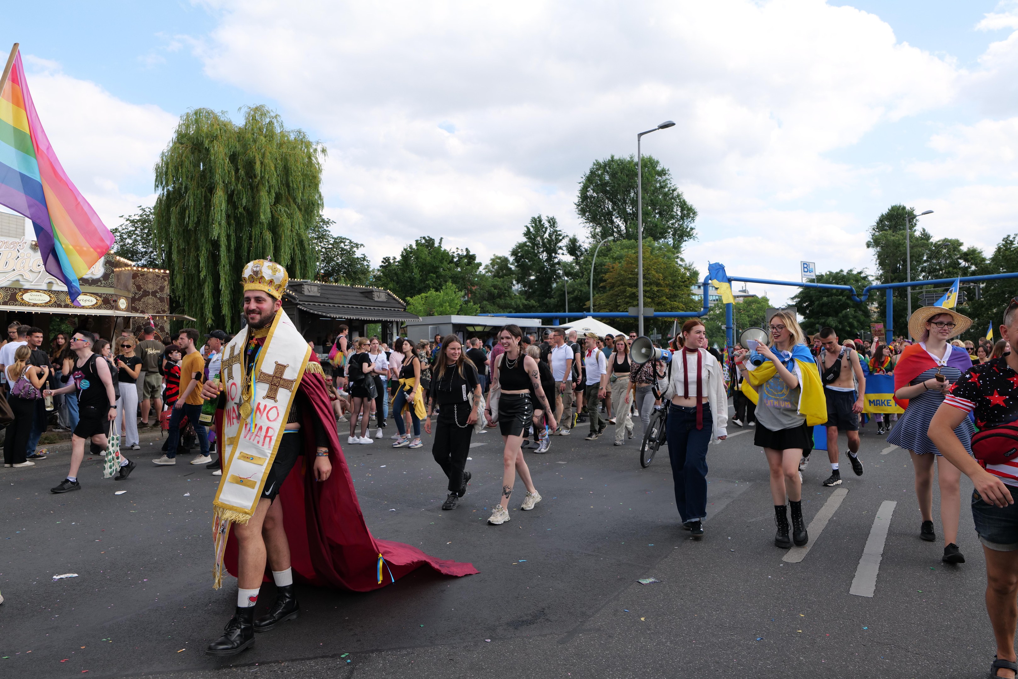 Eine Gruppe von Menschen marschiert bei der Pride Parade 2018 mit einer Regenbogenflagge und Musikinstrumenten, während im Hintergrund Laternenmasten, Bäume, Schuppen und ein bewölkter Himmel zu sehen sind.