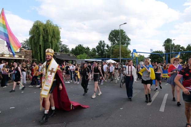 Eine Gruppe von Menschen marschiert bei der Pride Parade 2018 mit einer Regenbogenflagge und Musikinstrumenten, während im Hintergrund Laternenmasten, Bäume, Schuppen und ein bewölkter Himmel zu sehen sind.