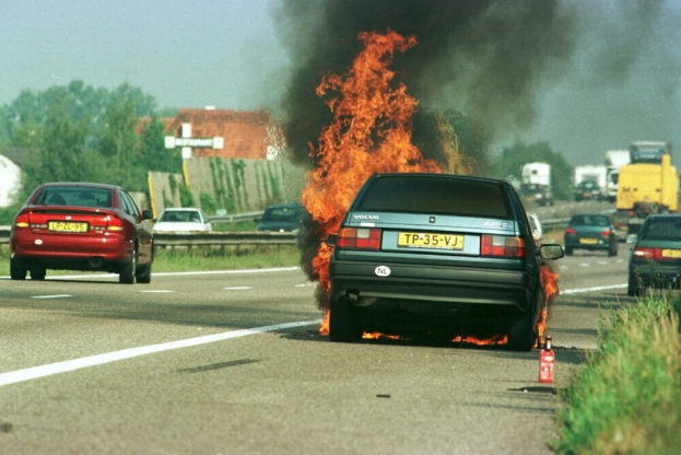 Ein Auto, das in Flammen steht, am Straßenrand umgeben von anderen Fahrzeugen, mit Bäumen, Gebäuden und einem klaren blauen Himmel im Hintergrund; Gras und ein Feuerlöscher sind auf der rechten Seite zu sehen.