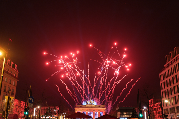 Eine belebte Stadtstraße bei Nacht am Silvesterabend in Berlin, mit Gebäuden, Bäumen, Laternenmasten, Verkehrszeichen, Zeltplanen, Menschen und einem prächtigen Feuerwerk, das den Himmel erhellt.