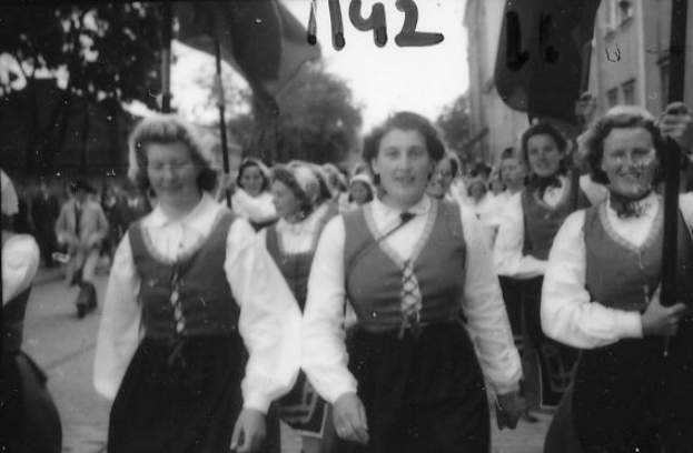 Eine Gruppe von Frauen in traditioneller bayrischer Tracht geht strahlend eine Straße entlang, umgeben von Bäumen und Gebäuden, auf einem schwarz-weißen Bild.