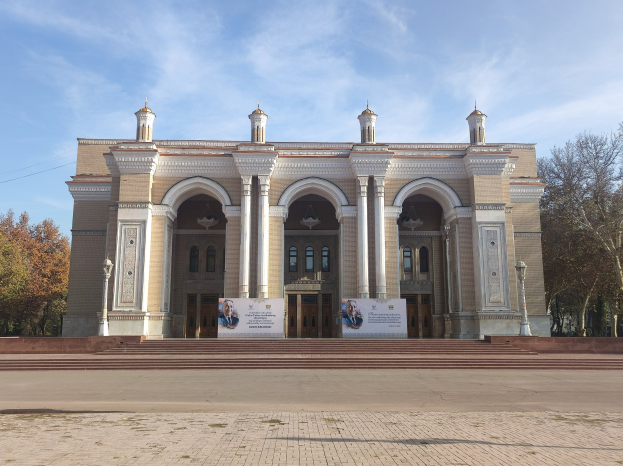 Großes Gebäude mit zahlreichen Fenstern, Säulen, Bögen und Treppen, identifiziert als Nationalmuseum der Russischen Föderation, mit Bannern, Laternenmasten, Bäumen und einem Draht sowie einem bewölkten Himmel im Hintergrund.