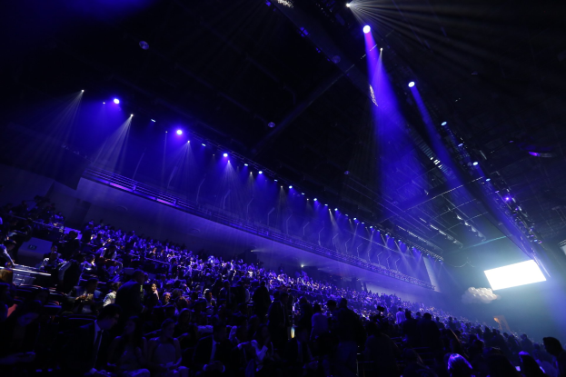 Große Menschenmenge in einem schwach beleuchteten Konzertsaal, der einer Bühne mit Deckenlichtern und einem Bildschirm auf der rechten Seite gegenüberliegt.