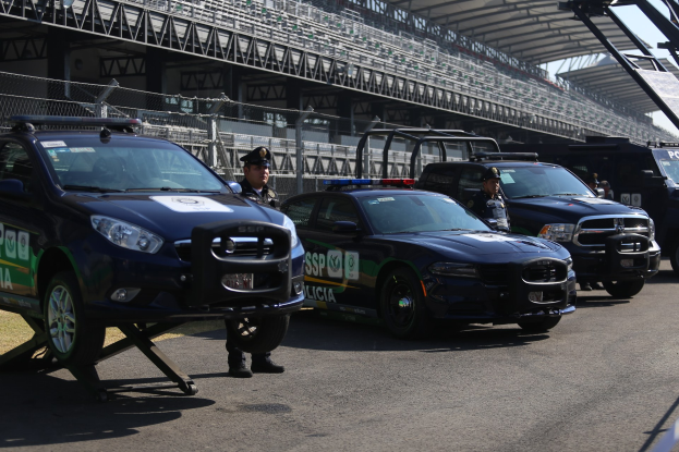 Polizeifahrzeuge vor einer Rennstrecke mit uniformierten Beamten, Stadion und Absperrung im Hintergrund.