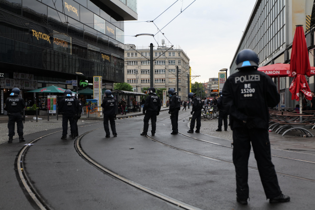 Eine Gruppe von Polizeibeamten in schwarzen Uniformen und Helmen steht auf einer von Glasfenstern gesäumten Straße, mit Bäumen, Strommasten und verstreuter Straßenmöblierung im Hintergrund unter einem klaren Himmel.