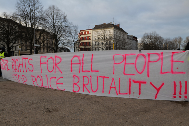 Eine Gruppe von Menschen hält ein Transparent mit der Aufschrift "Rechte für alle Menschen Stoppt Polizeigewalt" vor einem Straßenschild, einem Schild, Bäumen, Gebäuden mit Fenstern und einem bewölkten Himmel.