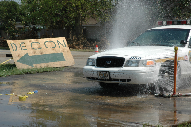 Ein Polizeiwagen fährt durch eine Pfütze, mit einem Schild, einer Verkehrsinsel und anderen Gegenständen auf dem Boden, mit Bäumen, Gebäuden und einem klaren blauen Himmel im Hintergrund.