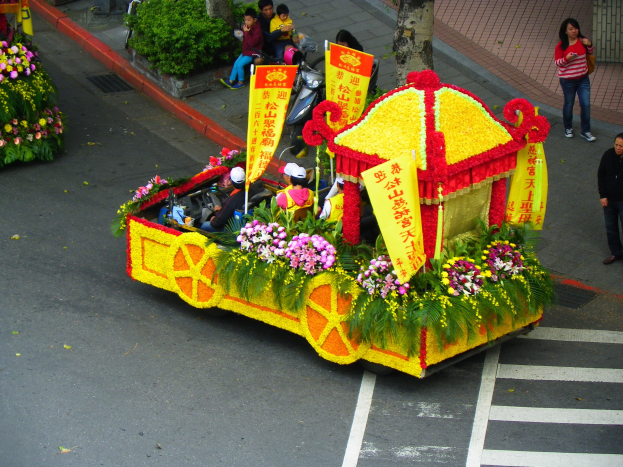 Schmuckvoll geschmückter Festwagen mit Blumen und Bannern in einem Umzug, auf dem Reiter sitzen, mit Zuschauern auf dem Gehweg rechts und Bäumen im Hintergrund.