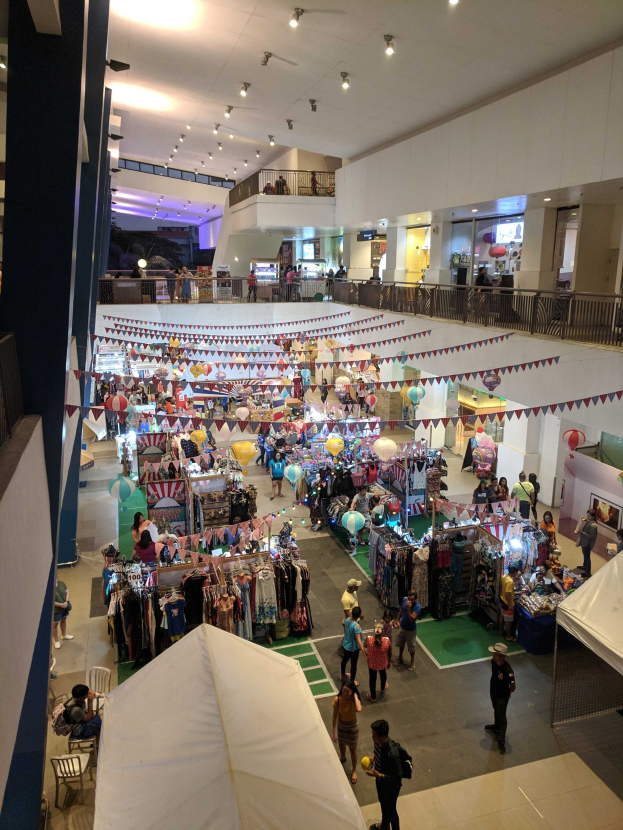 Ein volles Einkaufszentrum mit Kleiderständen, Dekorationsfahnen, Ballons, sitzenden und stehenden Menschen, Geländern, Säulen und Deckenleuchten, das an einen Weihnachtsmarkt im Mall of Asia erinnert.