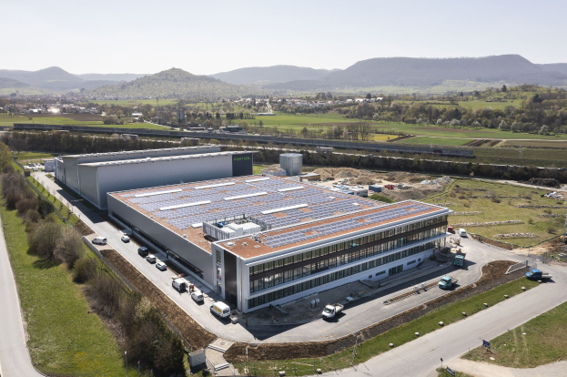 Luftaufnahme eines großen Gebäudes mit Solarmodulen auf dem Dach, umgeben von Fahrzeugen, Bäumen, Gras und Masten, vor Bergen und einem klaren blauen Himmel; das Gebäude ist Deutschlands erste solarbetriebene Fabrik.