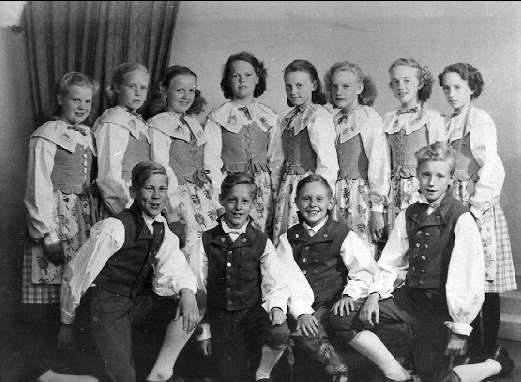 Schwarzes und weißes Foto einer Gruppe von Kindern, die vor einer Wand mit einem Vorhang posieren, beschriftet mit "1930er Kinder-Schule" unten.