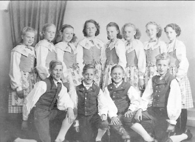 Schwarzes und weißes Foto einer Gruppe von Kindern, die vor einer Wand mit einem Vorhang posieren, beschriftet mit "1930er Kinder-Schule" unten.