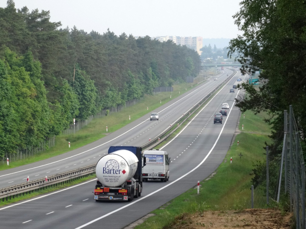 Ein großer Lkw fährt auf einer von Bäumen gesäumten Autobahn, mit einem Zaun und Gras auf der rechten Seite und Gebäuden im Hintergrund unter einem klaren blauen Himmel.