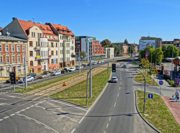 Stadtstraße mit Autos, hohen Gebäuden, Verkehrsampeln, Schildern, Fußgängern, Bäumen und Gras unter einem Himmel.
