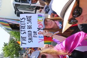 Eine Gruppe von Menschen steht vor einem Lastwagen und hält ein Schild hoch, auf dem "Die Straßen sollten sicher sein" steht, einige tragen Mützen und Schutzbrillen, andere halten Schilder, vor einem Hintergrund aus Bäumen, Gebäuden und einem klaren blauen Himmel.