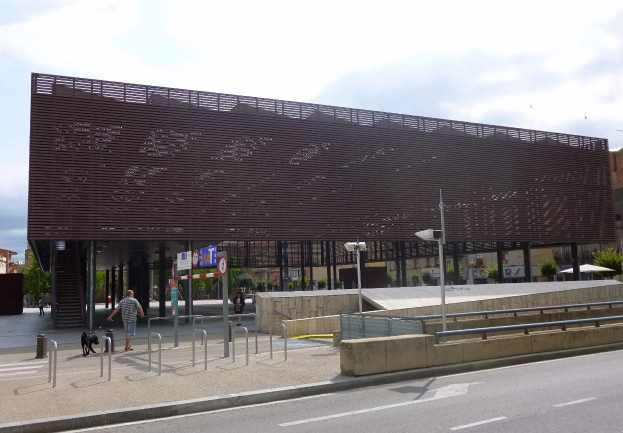 Großes modernes Gebäude mit zahlreichen Fenstern, das als neuer Eingang zum Stadtzentrum dient, umgeben von städtischer Infrastruktur einschließlich Straßenlaternen, Fahrzeugen, Fußgängern und Bäumen unter einem bewölkten Himmel.