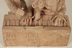 Eine steinerne Skulptur eines gekrönten Mannes in einem langen Gewand, der mit geschlossenen Augen auf einem Klavier spielt.