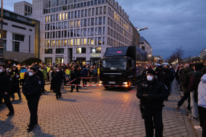 Eine Gruppe von Menschen steht vor einem Lkw auf einer Straße, umgeben von Gebäuden, Laternenmästen, Bäumen und einem bewölktem Himmel, wobei einige Mötzen und Masken tragen und ein Band mit einem Pfahl im Vordergrund ist.