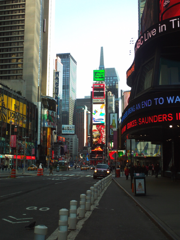 Eine belebte Stadtstraße mit hohen Gebäuden, Fahrzeugen, Fußgängern, Pollern und an Gebäuden angebrachten Schildern mit Text und Bildern.