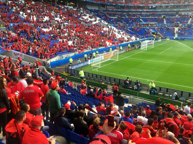 Eine große Menge Menschen in einem Stadion schaut einem Fußballspiel zu, mit Zuschauern auf Sitzen und Stehplätzen, Schildern, einem Torpfosten und Deckenlampen unter einer Dachkonstruktion.