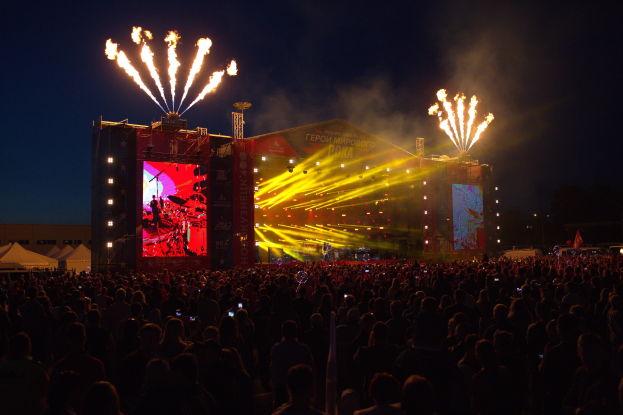 Eine große Menge bei einem Musikfestival schaut auf eine hell erleuchtete Bühne mit Zelten links und Feuerwerk am dunklen Himmel dahinter.