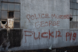 Ein Gebäude mit Graffiti, das "Polizei Mord George Fuck 12 P19" auf der Seite trägt, mit Fenstern und verschiedenen Gegenständen davor.