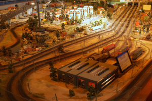 Eine detaillierte Miniatur-Modellbahnanlage in einem Museum, die Gebäude, Bäume, Eisenbahnschienen, Fahrzeuge, Masten, Lichter und andere Gegenstände zeigt, die eine belebte Stadtlandschaft nachahmen.