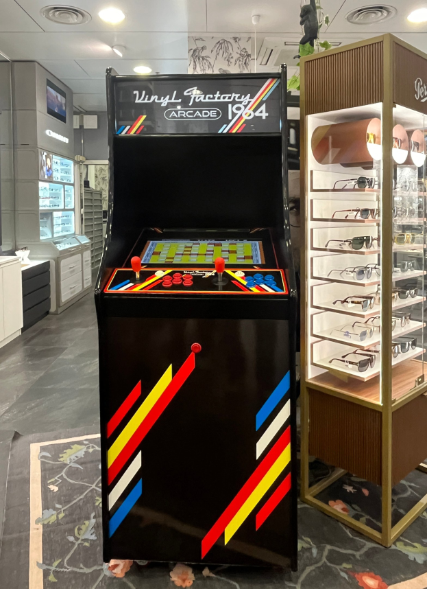 Ein Geschäft mit einem zentralen Vintage-Arcade-Automaten, einem Gestell mit Gläsern auf der rechten Seite und Hintergrund-Elementen wie Schränken, einem Fernseher und anderen Gegenständen, mit Deckenleuchten, die eine retro-mäßige Atmosphäre schaffen.