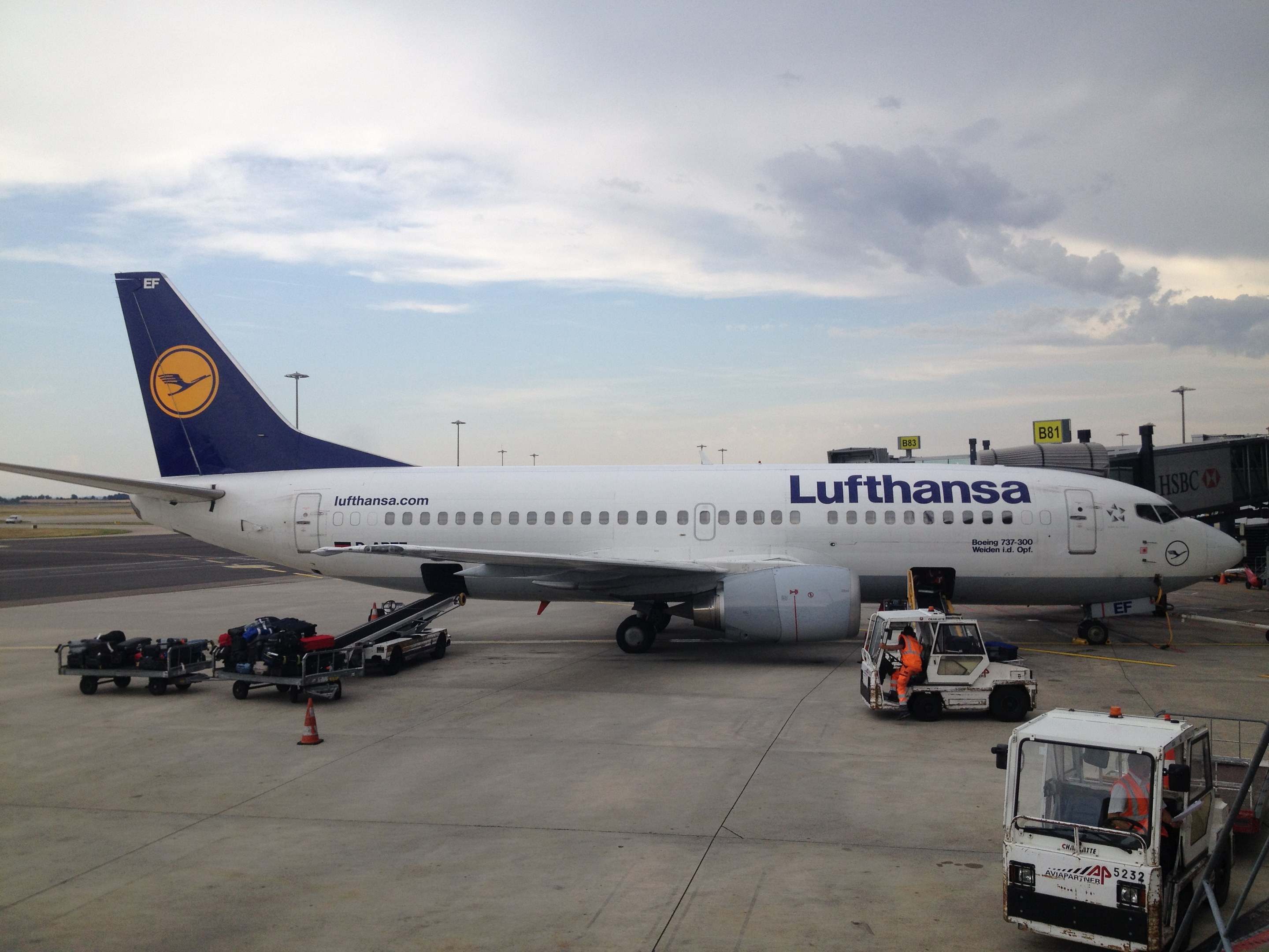 Airbus A320-200 der Lufthansa auf dem Frankfurt Airport geparkt mit Bodenfahrzeugen, Verkehrskegeln und Informationsschildern im Hintergrund bei bewölktem Himmel.