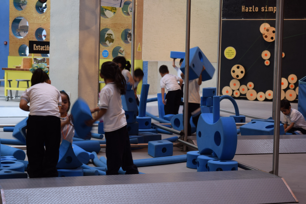 Kinder beim Spielen in einer Turnhalle, einige halten Gegenstände, mit einer Texttafel an der Wand und Stangen auf der rechten Seite.