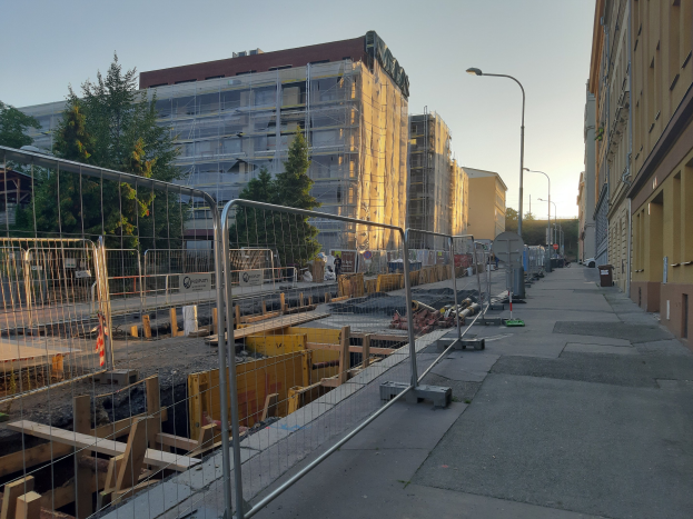 Stadtstraße mit Baustelle umgeben von Gebäuden, Straßeninfrastruktur, Fahrzeugen, Bäumen und einem klaren blauen Himmel.