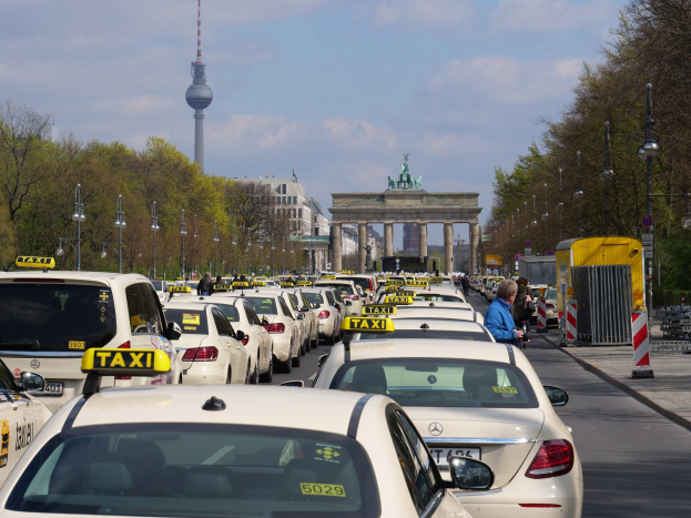 Eine belebte Straße in Berlin mit zahlreichen geparkten Taxis, Fußgängern auf dem Gehweg, Laternenpfählen, Bäumen, Gebäuden, fernen Statuen unter einem Bogen und einem Turm unter einem bewölkten Himmel.