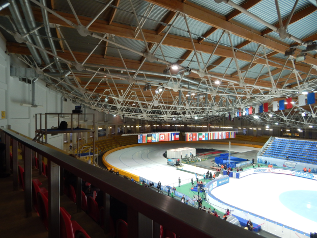 Großes Innen-Eisstadion voller Menschen, umgeben von Sitzplätzen, Geländern, Tafeln, Beleuchtung und strukturellen Elementen, mit einer Wand im Hintergrund.