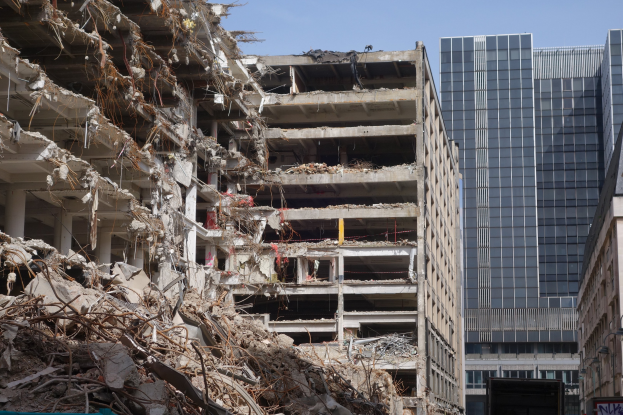 Ein großes Gebäude wird in der Innenstadt abgerissen, umgeben von intakten Gebäuden, Straßeninfrastruktur, Fahrzeugen, Bäumen und einem klaren Himmel.