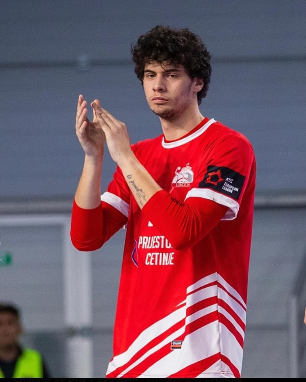 Ein Mann in einem roten und weißen Hemd, der in der Vordergrund klatscht, mit einer weiteren Person und einer Wand im Hintergrund und einem Text, der unten 'Copenhagen Futsal Club - Foto 1' lautet.