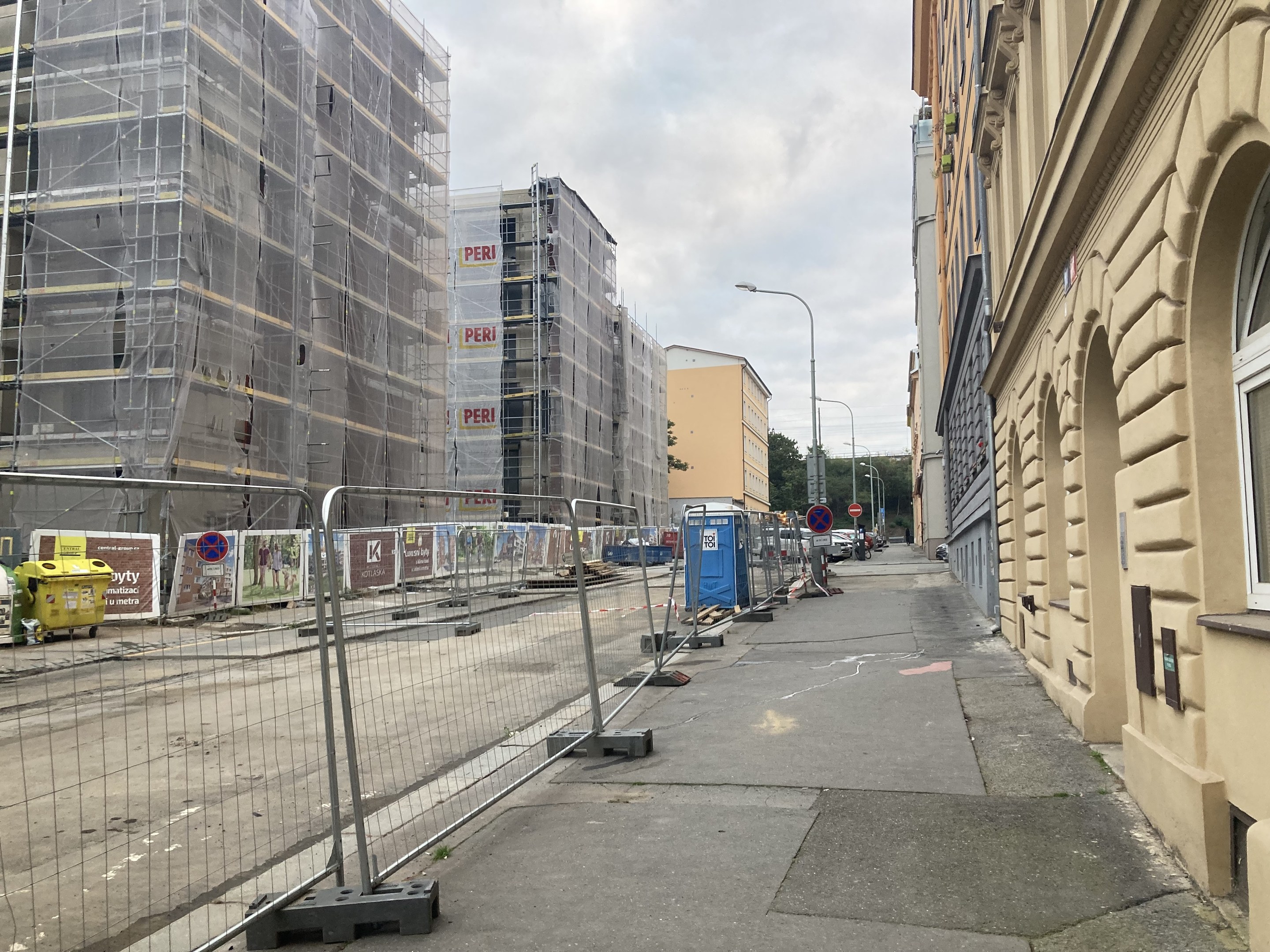Stadtstraße mit Gebäuden, Fahrzeugen, Straßeninfrastruktur, Bäumen und einer Baustelle mit Gerüsten im Vordergrund.
