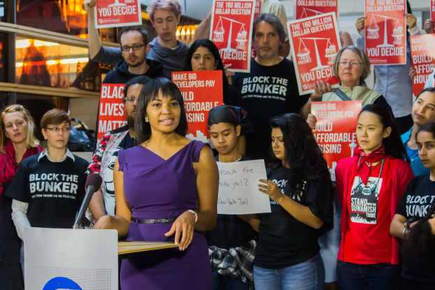 Eine Black-Lives-Matter-Aktivistin spricht vor einem Publikum mit Mikrofon und Schildern, mit Lichtern und anderen Gegenständen im Hintergrund.