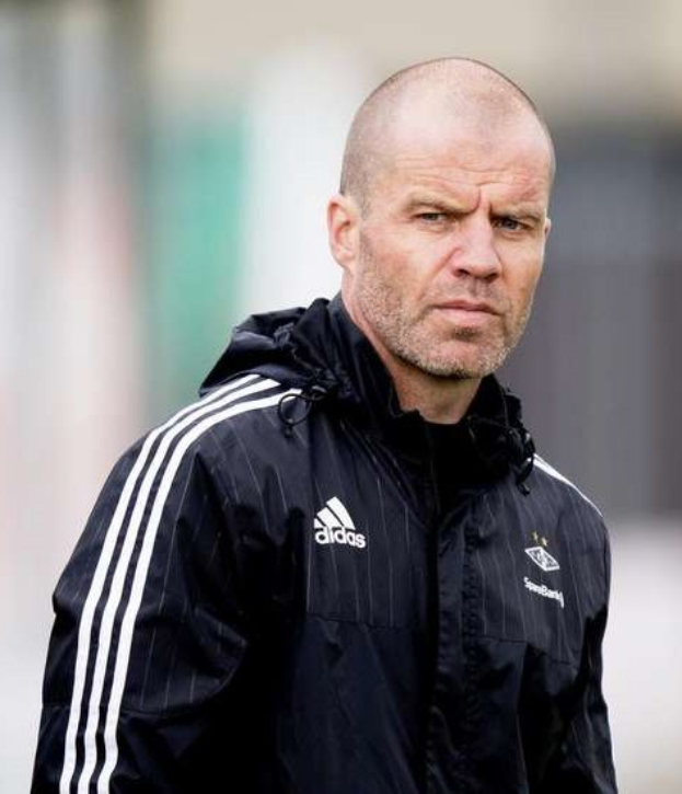 Mann in einem blauen Jackett mit weißen Streifen und weißem Kragen, der auf einem Fußballfeld steht und die Hände verschränkt hält.