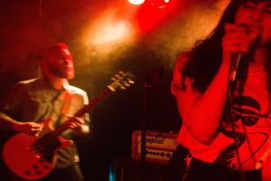 Eine Frau singt in ein Mikrofon auf der Bühne, während ein Mann im Hintergrund Gitarre spielt, mit Deckenleuchten, die die lebendige Kulisse erhellen.