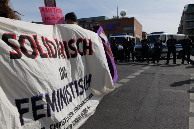 Gruppe von Menschen marschiert auf einer Straße mit einem 'Solidarität und Feminismus'-Schild, während geparkte Autos und Gebäude im Hintergrund zu sehen sind, unter einem klaren blauen Himmel.