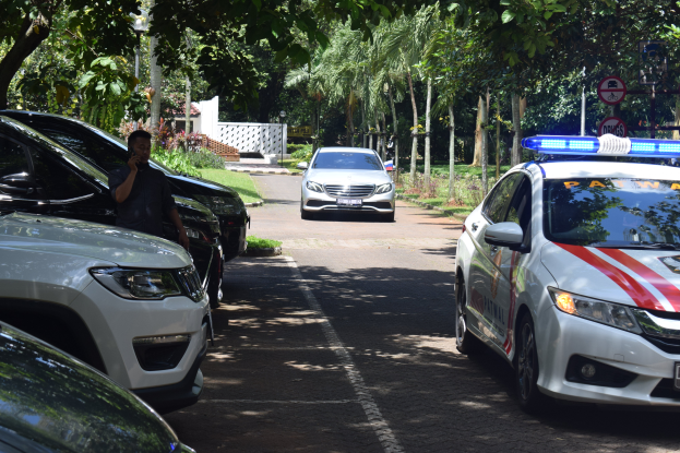 Ein Polizeiwagen steht auf der Seite einer Straße neben anderen Fahrzeugen, wobei eine Person auf der linken Seite ein Mobiltelefon hält und im Hintergrund Bäume, Schilder, Pfosten, Pflanzen, Gras und eine Wand zu sehen sind.